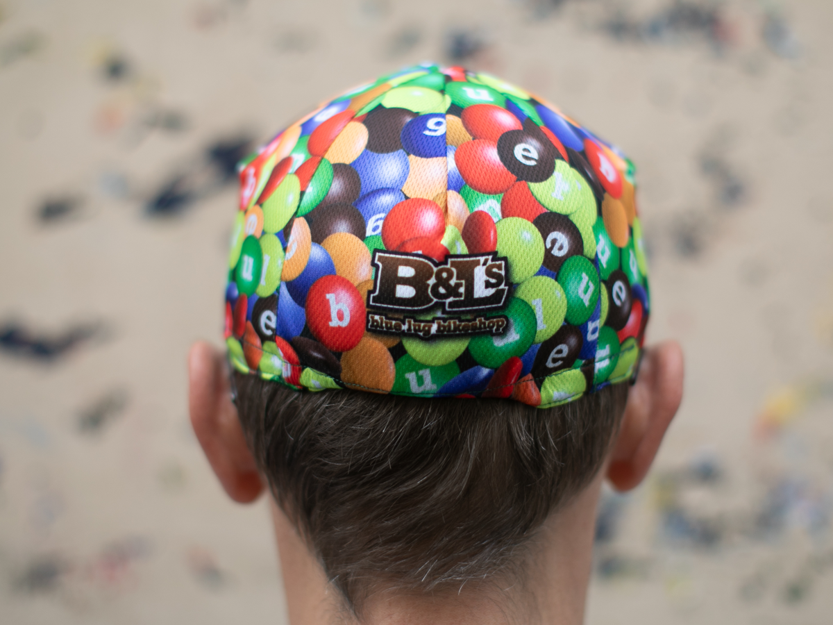 Vue arrière de la casquette de cyclisme Blue Lug coloris Chocolate/Poly, avec le patch logo "B&L's Blue Lug Bikeshop" stylisé en parodie de la marque M&M's.