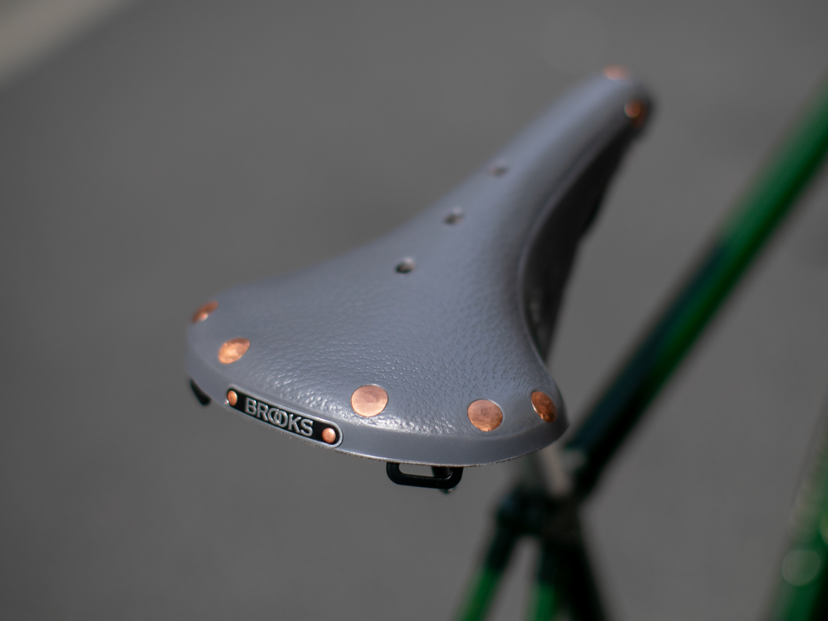Selle Brooks x Rivendell B17 coloris gris — vue trois quarts avant sur cadre vert, montrant toute la surface en cuir pebble avec rivets cuivre des deux côtés et badge Brooks au nez