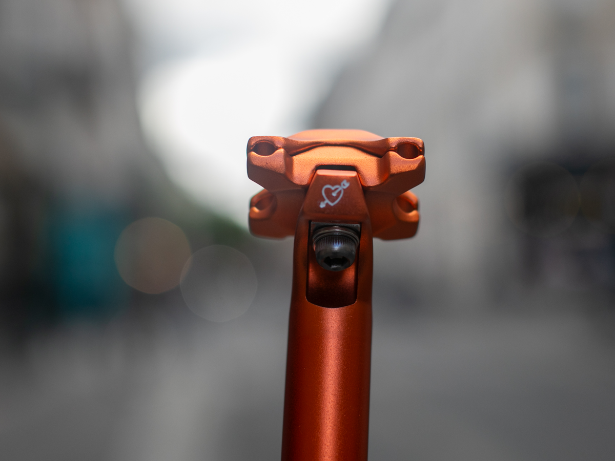 SimWorks Beatnik Sunrise seat post — front view of the clamp showing the hand-engraved cupid heart detail and saddle bolt in Sunrise orange