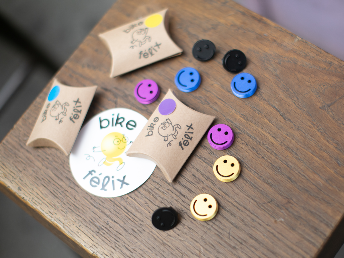 Bike Felix Smiley aluminium crankset caps in black, blue, purple and yellow, displayed alongside recyclable kraft cardboard packaging and Bike Felix sticker