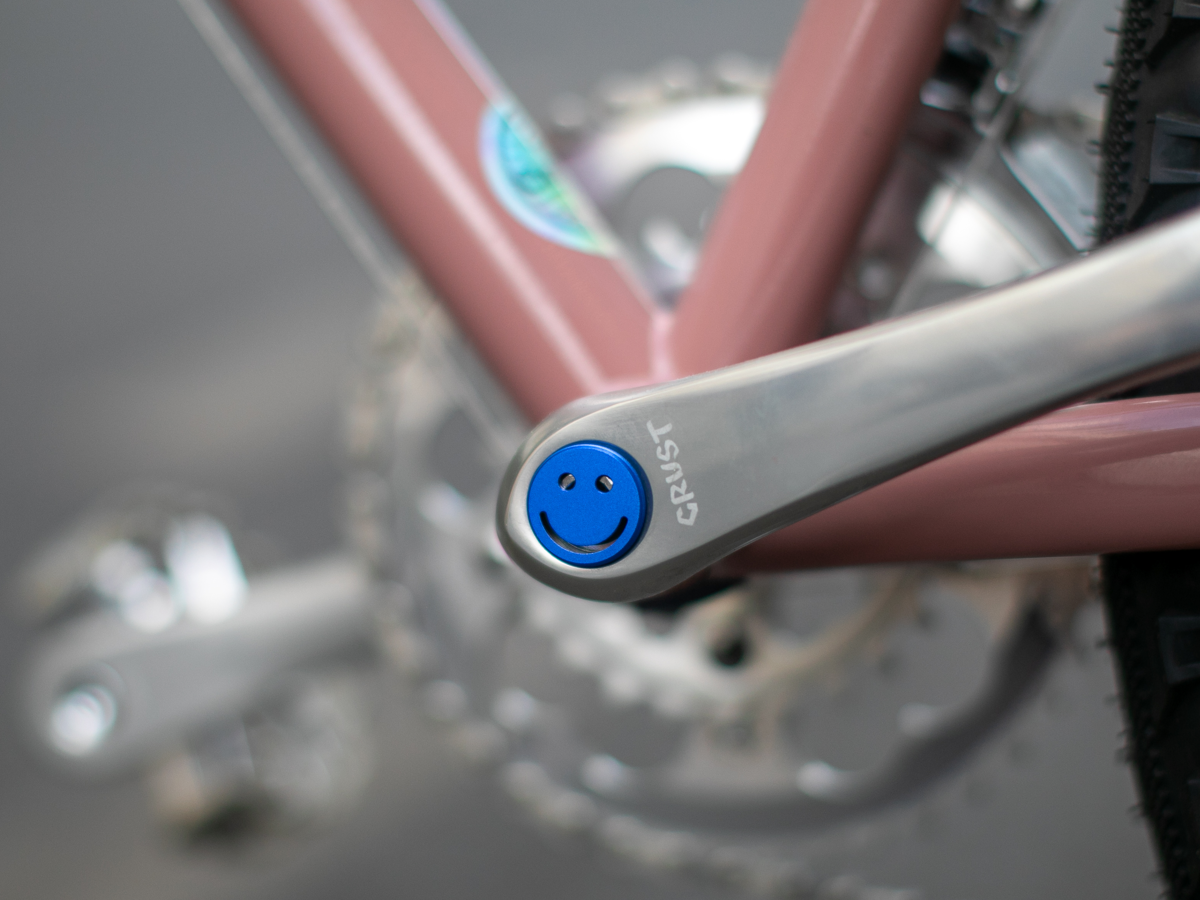 Blue Bike Felix Smiley crankset cap installed on a silver Crust crankarm on a pink bicycle