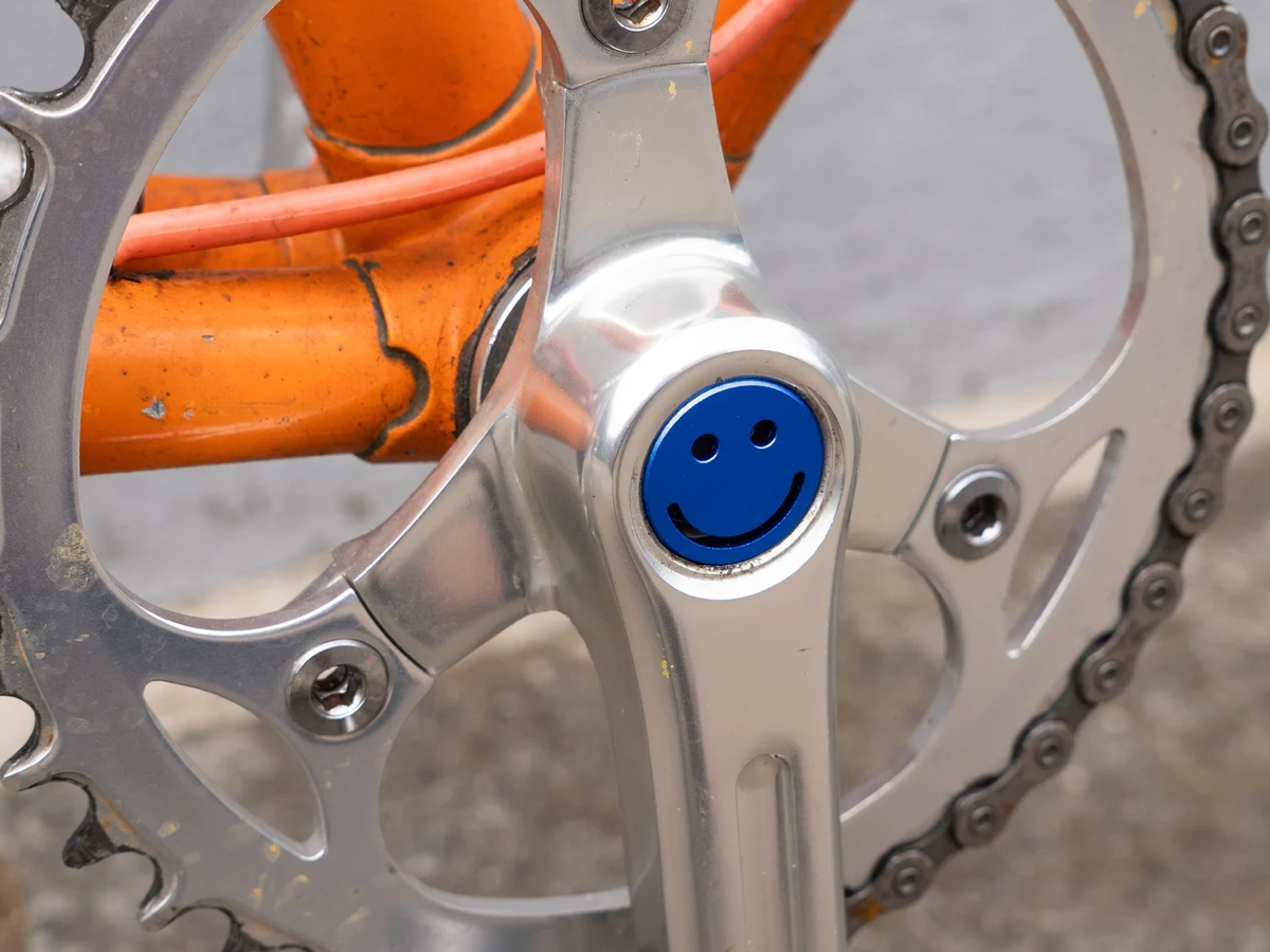 Blue Bike Felix Smiley aluminium crankset cap fitted to a silver double chainring crankset on an orange bike
