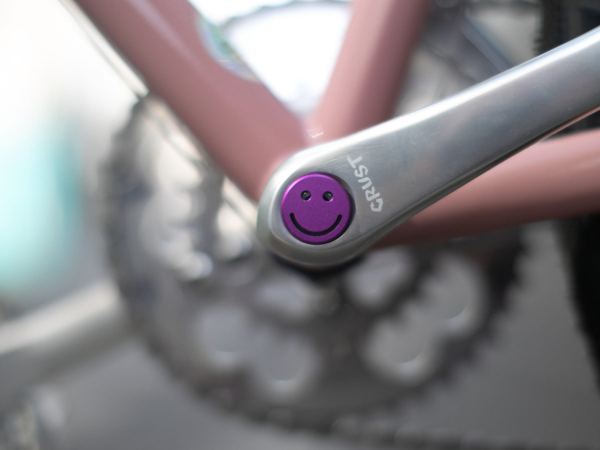 Purple Bike Felix Smiley crankset cap installed on a silver Crust crankarm on a pink bicycle
