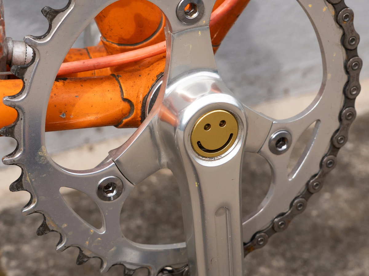 Yellow gold Bike Felix Smiley aluminium crankset cap fitted to a silver crankset on an orange bike