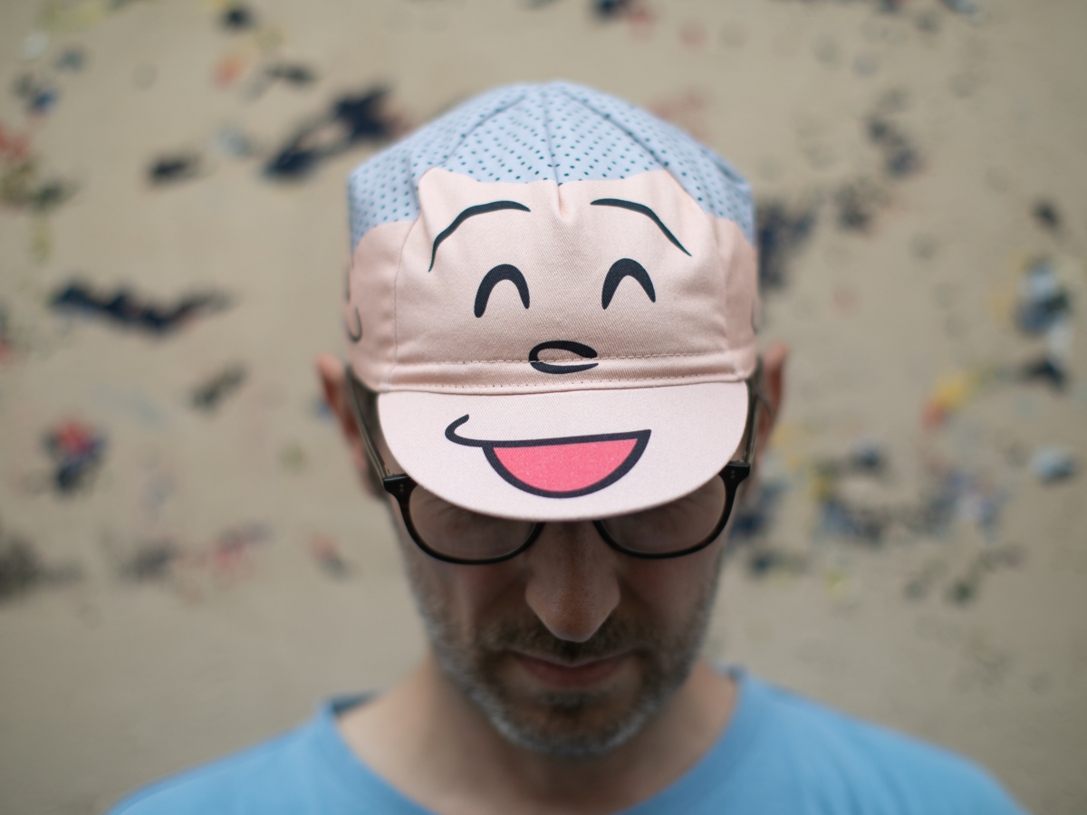 Blue Lug Good Boy Cycle Cap worn brim-down, front view. The front panels feature a large cartoon smiley face in pink with squinting eyes and an open smile, on a light blue polka dot crown.
