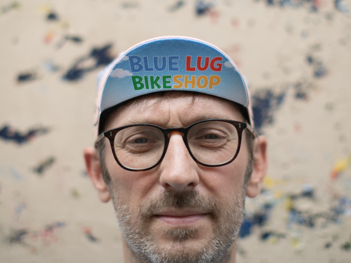 Blue Lug Good Boy Cycle Cap worn brim-up, front view. The light blue brim displays "Blue Lug Bikeshop" in bold multicolour lettering on a sky and clouds graphic.