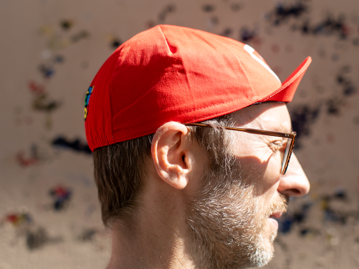 Profil de la casquette de cyclisme Blue Lug Jump and Dash rouge, montrant la visière pointue caractéristique et un aperçu du logo multicolore sur le panneau arrière.