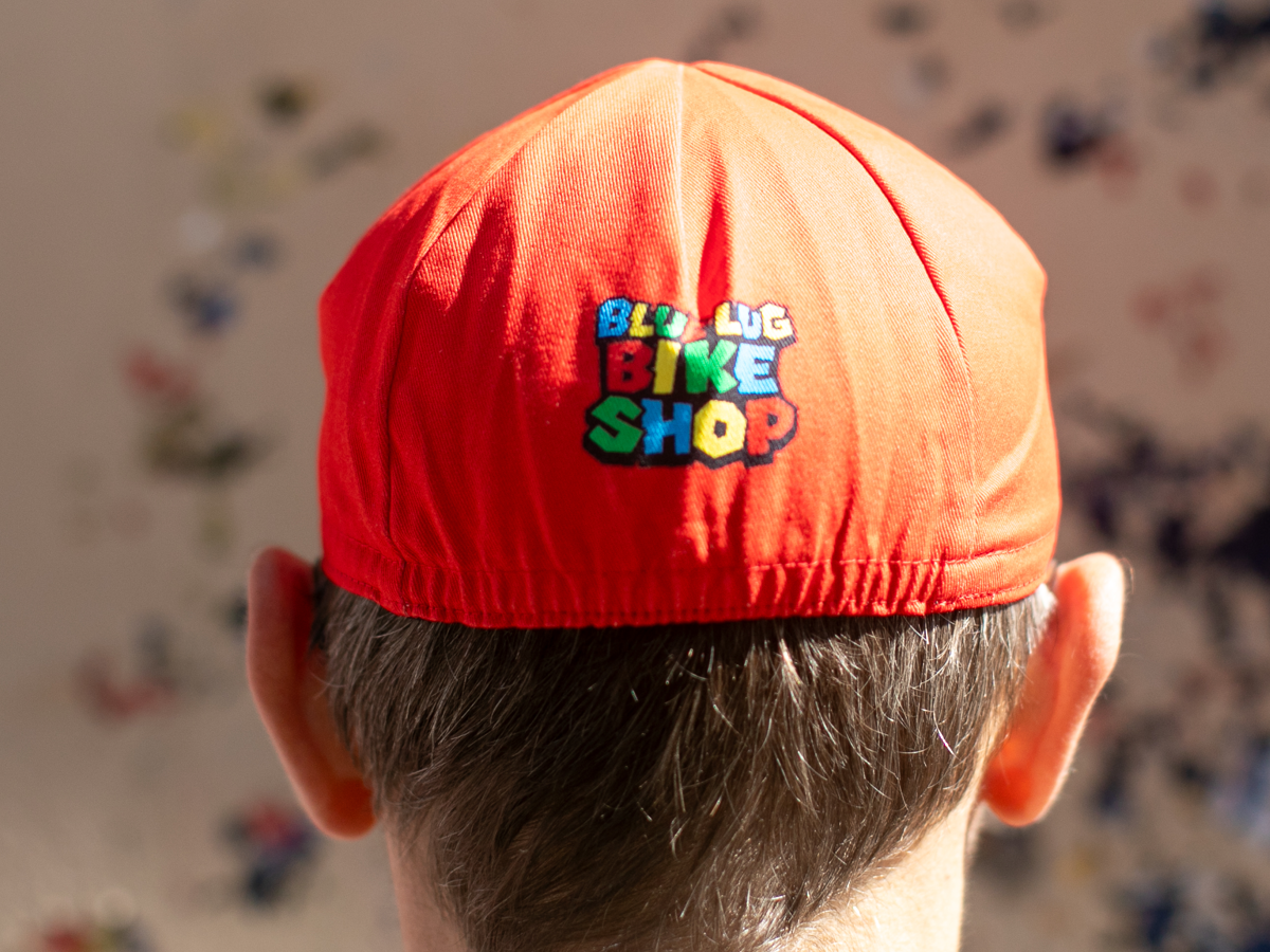 Vue arrière de la casquette de cyclisme Blue Lug Jump and Dash rouge, avec le logo "Blue Lug Bike Shop" en lettres multicolores style jeu vidéo sur le panneau arrière.