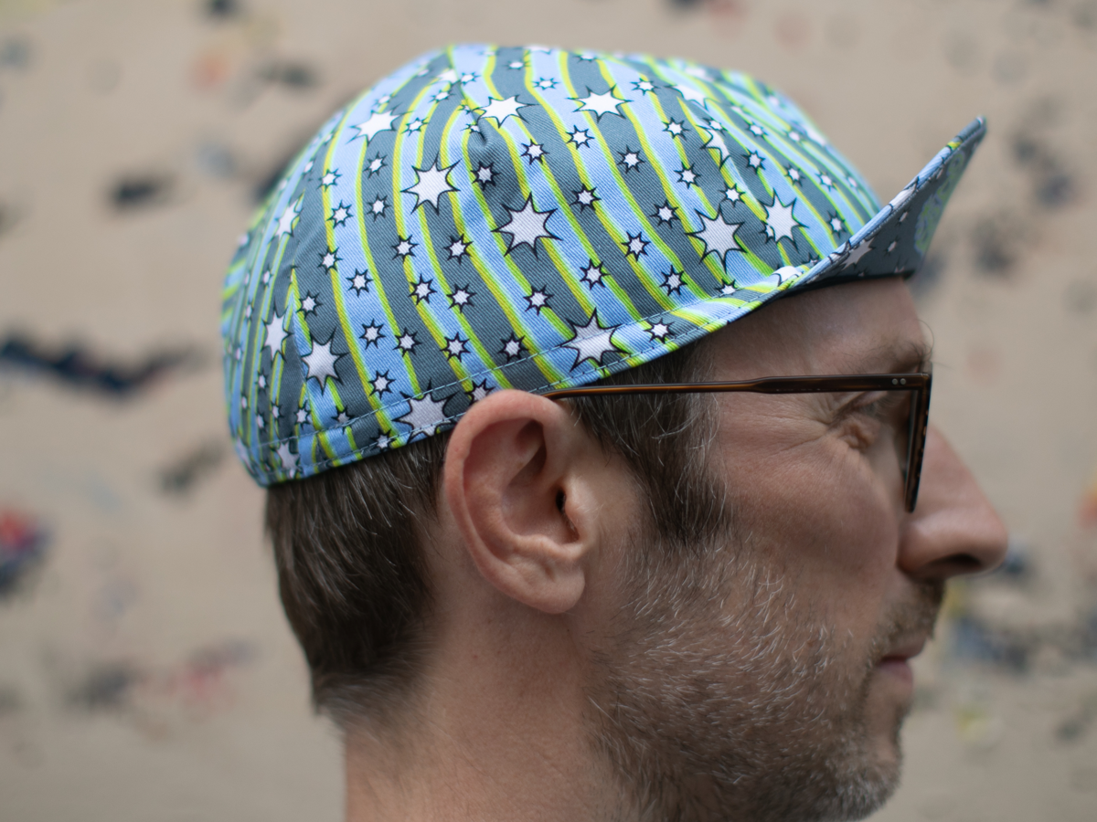 Side profile of the Blue Lug Cycle Cap in Dreamer, showing the swirling stripe and star print in blue, grey and neon yellow-green, and the classic pointed cycling cap brim.