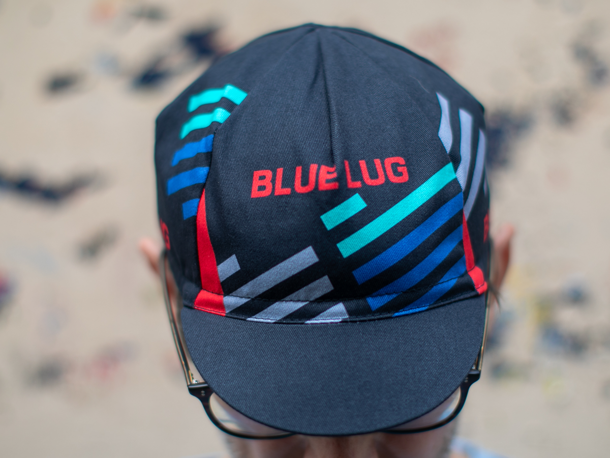 Blue Lug Original Cycle Cap in black, seen from above. The geometric stripe pattern in cyan, blue, grey and red, with "BLUE LUG" in red text, is visible across the panels.