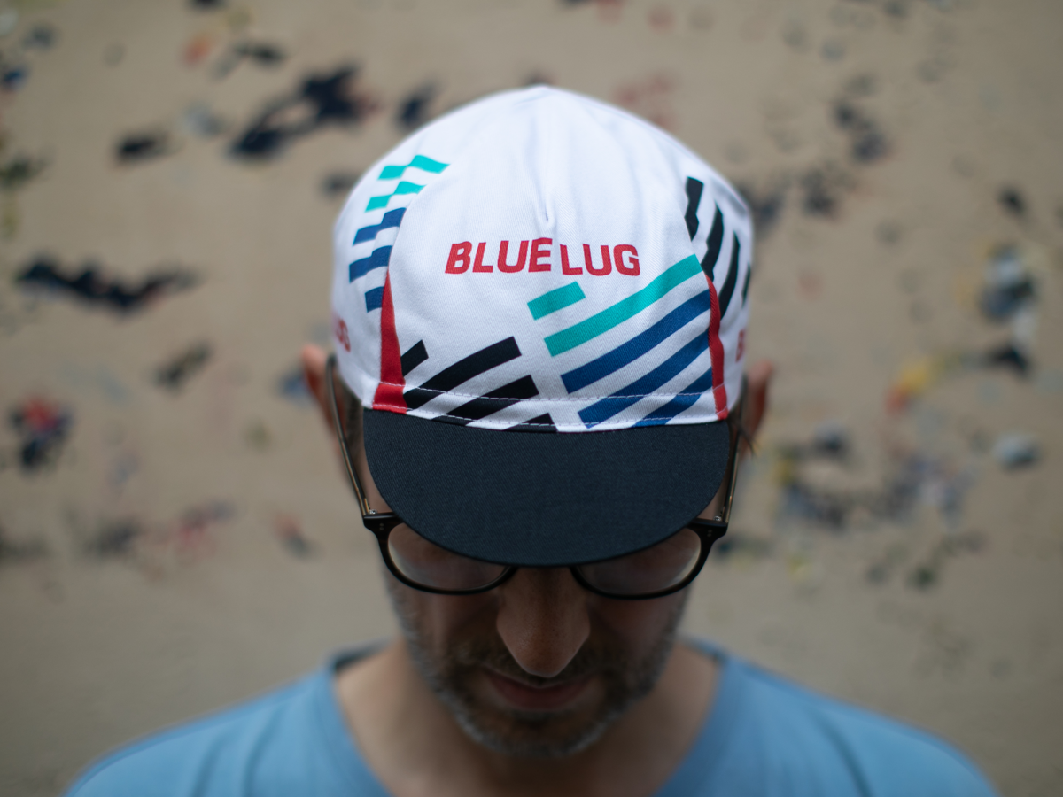 Blue Lug Original Cycle Cap in white, seen from above. The geometric stripe pattern in cyan, blue, black and red with "BLUE LUG" in red text covers the panels.