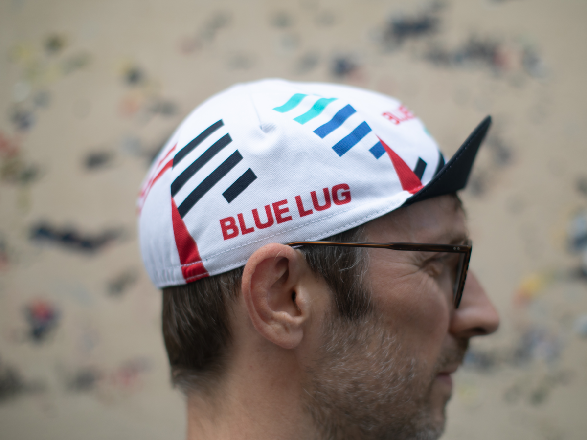 Side profile of the Blue Lug Original Cycle Cap in white, showing the geometric stripe design, "BLUE LUG" text in red, and the classic pointed brim.
