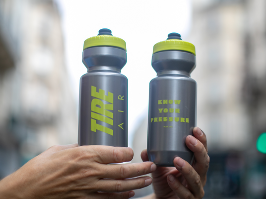 Blue Lug Tire Air water bottle, both sides shown. Left side reads "TIRE AIR" in bold neon yellow lettering; right side reads "KNOW YOUR PRESSURE" with the Blue Lug logo below. Grey body with neon yellow cap.