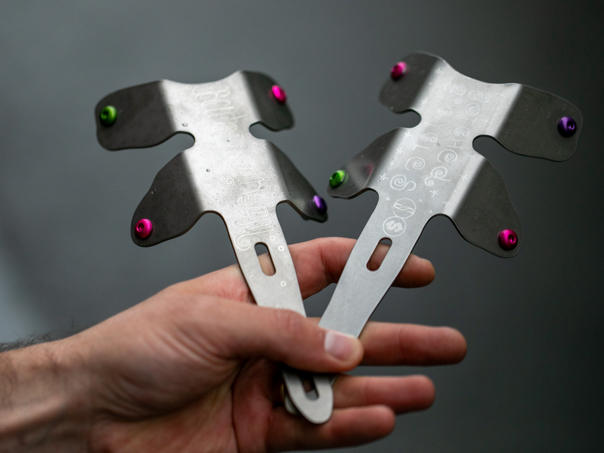 Une main tenant deux adaptateurs Botched Bikes Titanium Dragonfly en éventail, montrant le profil en titane formé avec les ailes de fixation courbées et la gravure laser Botched Bikes sur la surface. Boulons anodisés en rose, vert et violet.