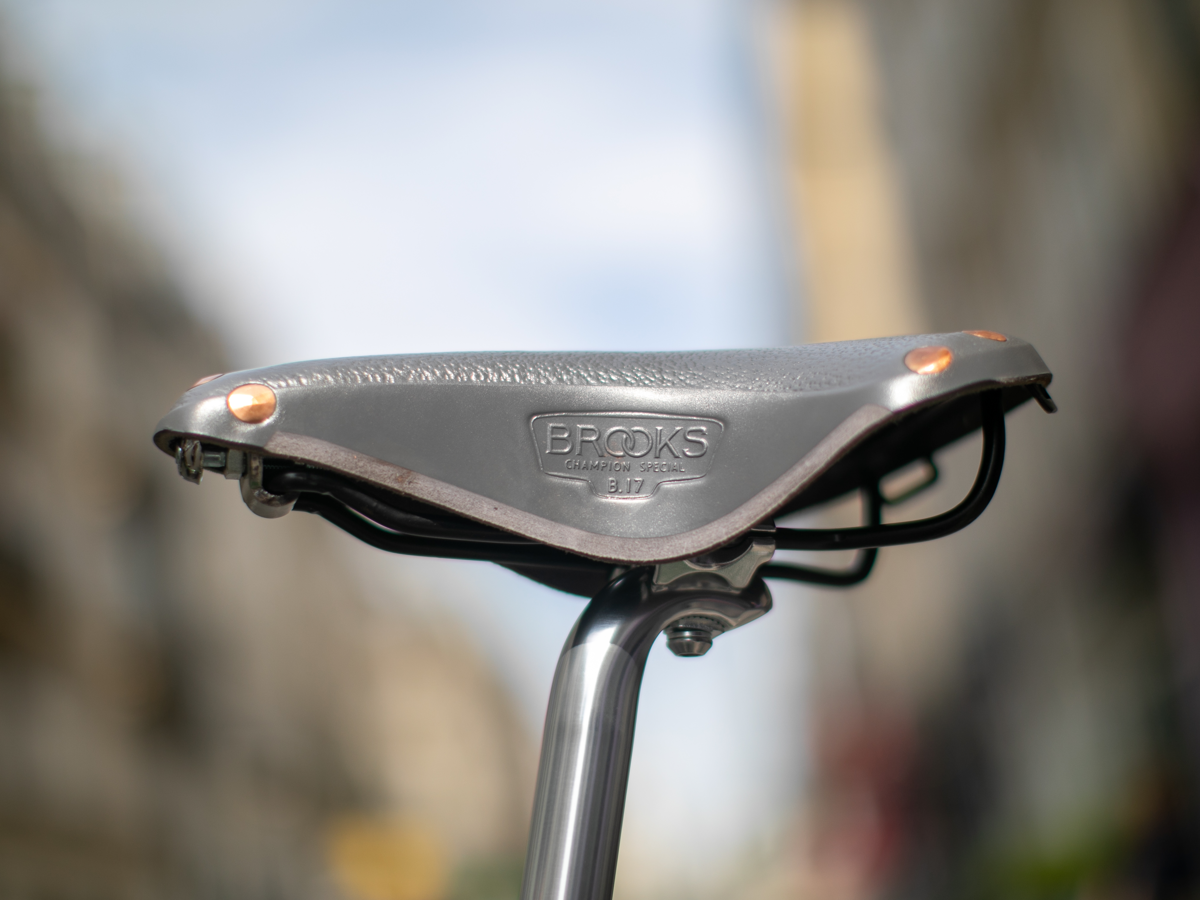 Selle Brooks x Rivendell B17 coloris gris — vue de côté sur tige de selle argentée, avec dessus en cuir pebble, rivets cuivre au nez, flanc inférieur sculpté et estampille "Brooks Champion Special B17"