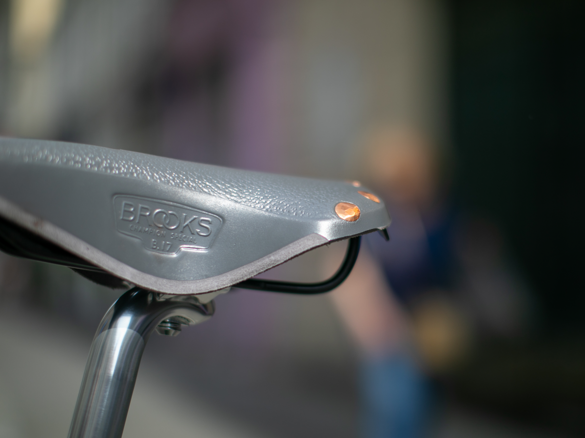 Selle Brooks x Rivendell B17 coloris gris — vue latérale arrière sur tige de selle argentée, avec dessus pebble, rivets cuivre à la queue, rails acier noir et estampille "Brooks Champion Special B17", fond urbain