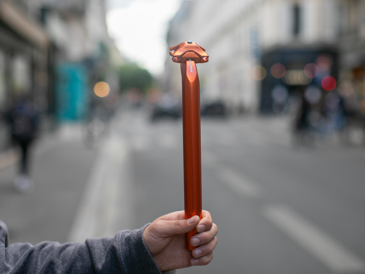 SimWorks Beatnik Sunrise seat post in Sunrise orange — full view of the 300mm forged aluminum post with saddle clamp, held up against a blurred Paris street