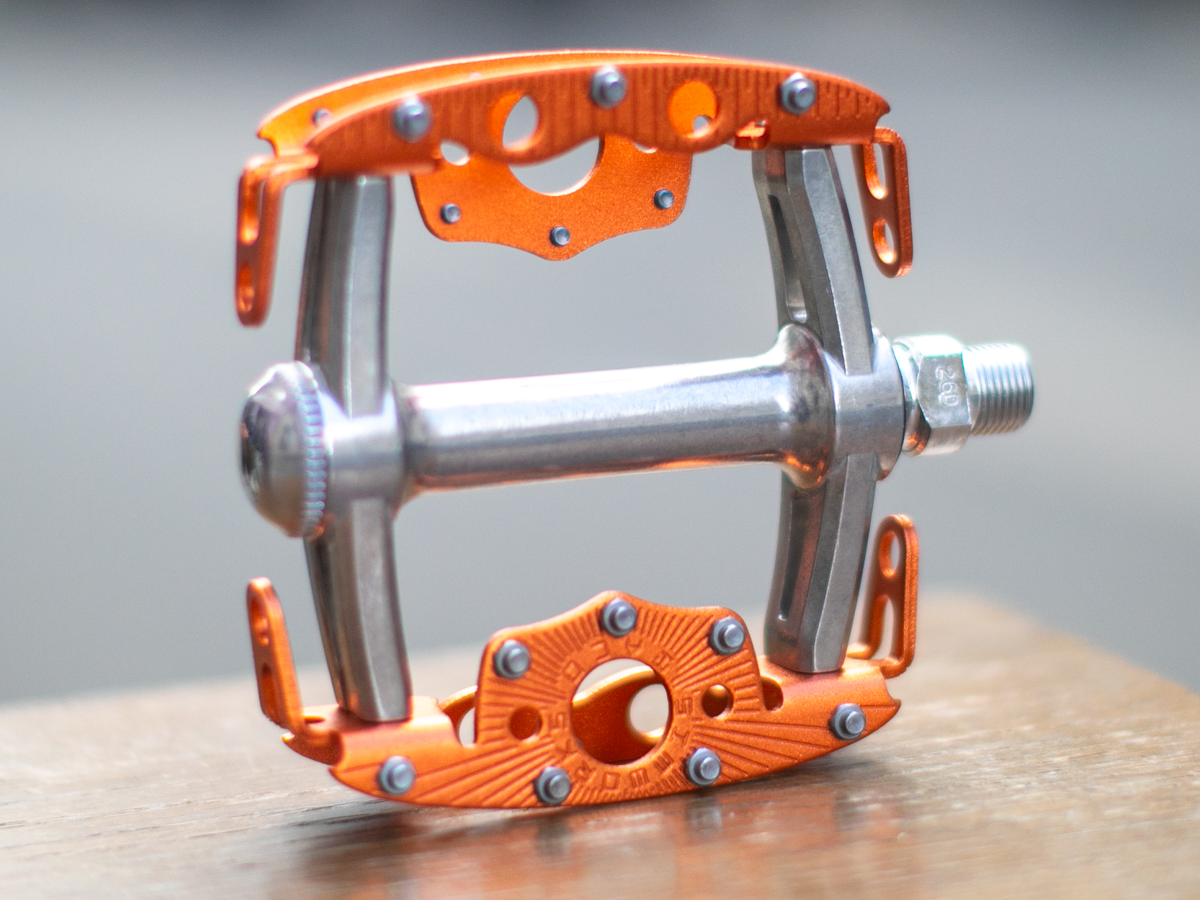 SimWorks Taco Pedal in Sunrise orange — side profile showing the open cage structure, grip studs, silver axle with cup-and-cone bearing adjustment, and SimWorks sunburst engraving on the platform