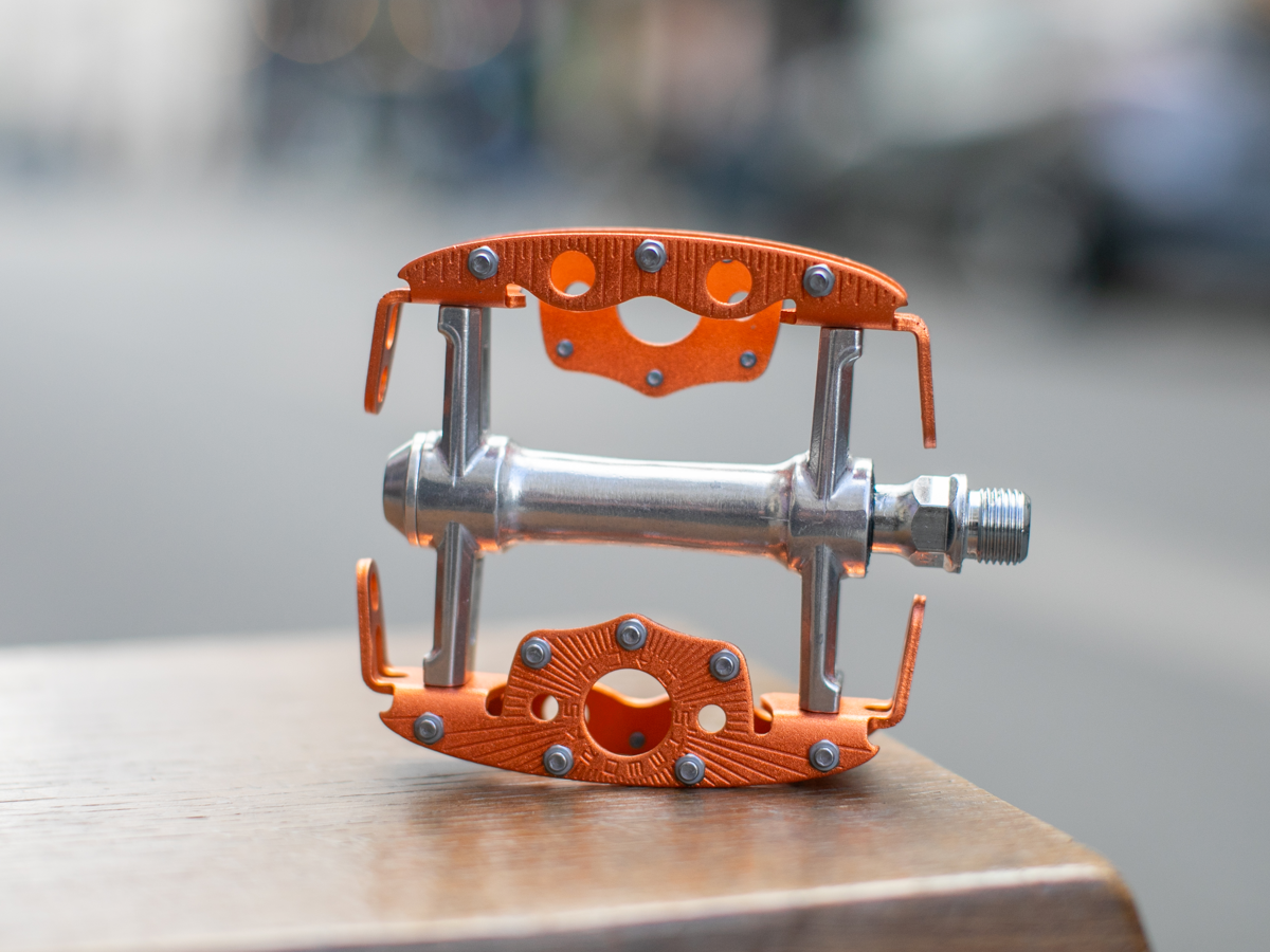 SimWorks Taquito Pedal in Sunrise orange — side profile on a wooden surface showing the compact open cage structure, grip studs, CrMo axle and SimWorks sunburst engraving on the platform