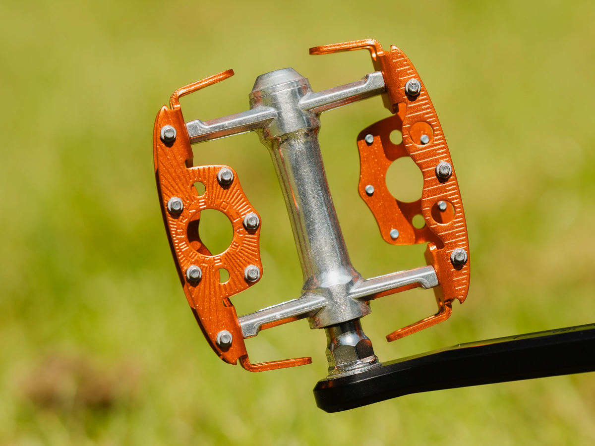 SimWorks Taquito Pedal in Sunrise orange — front view installed on a black crank arm, showing the concave platform, open cage sides and grip studs against a green background